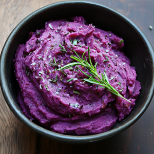 Vibrant purple sweet potato mash garnished with fresh herbs, served in a white bowl on a rustic wooden table.