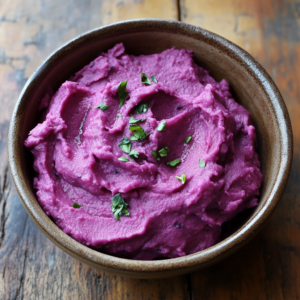 Vibrant purple sweet potato mash garnished with fresh herbs, served in a white bowl on a rustic wooden table.